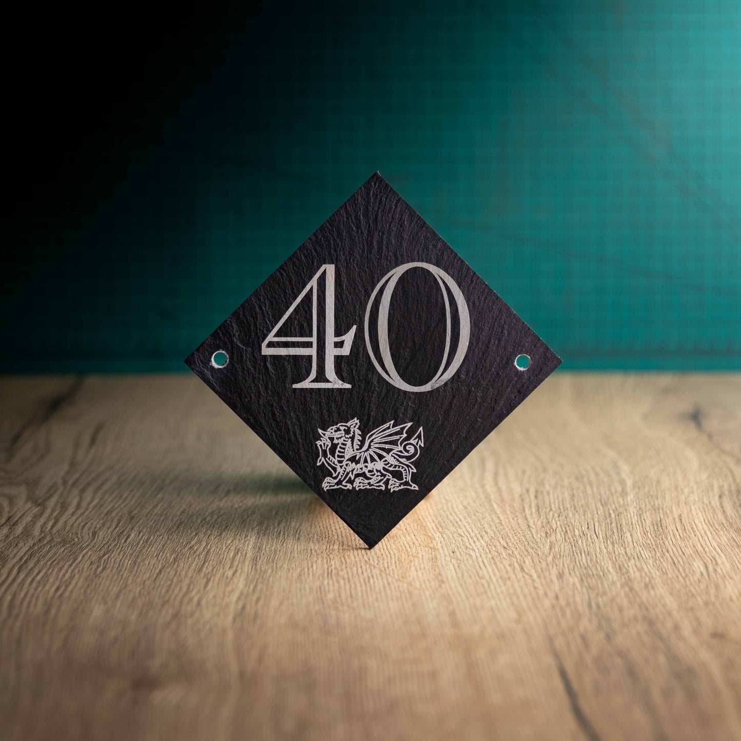 A laser-engraved diamond slate house sign on a wooden surface and teal background. Diamond 4.5" x 4.5"