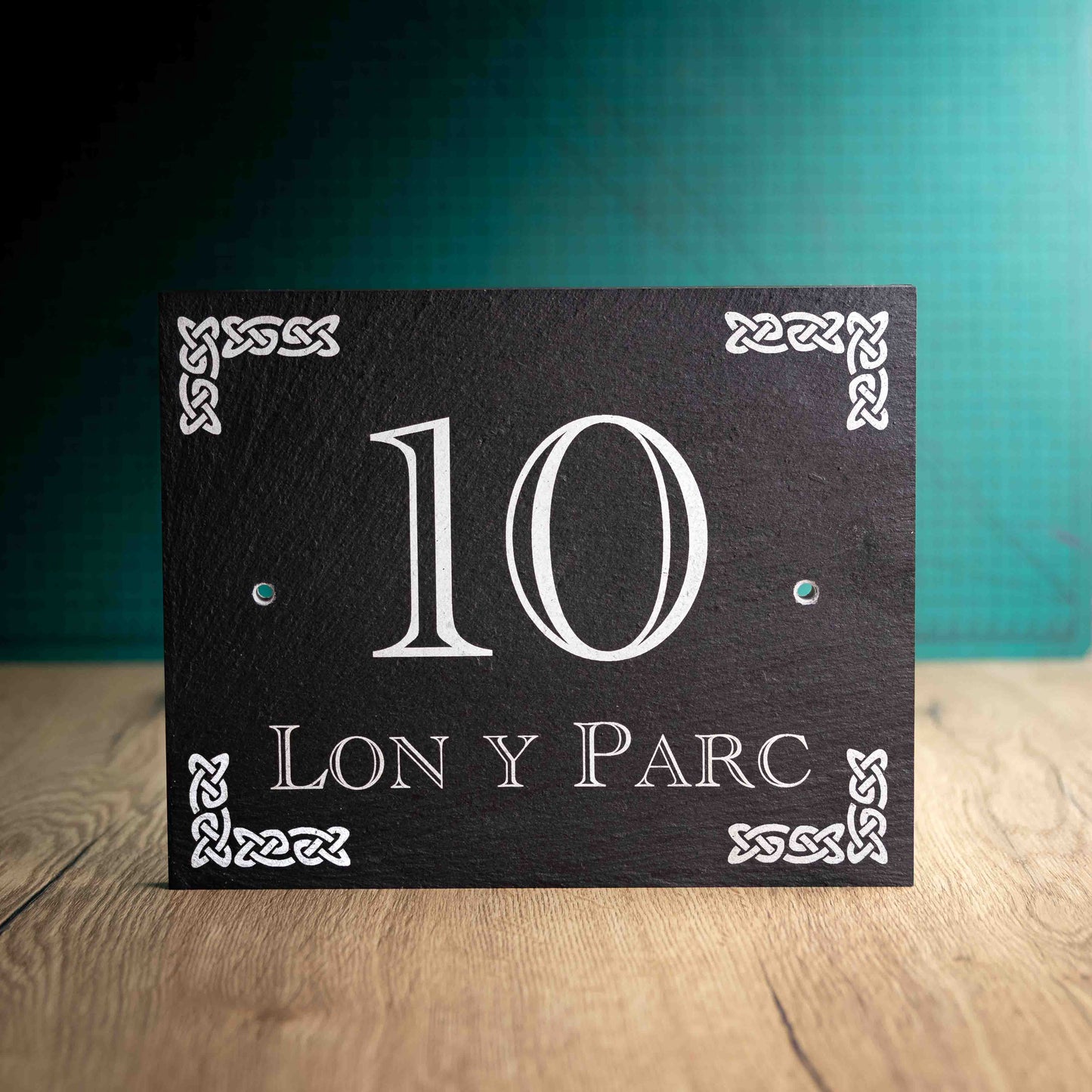 A laser-engraved slate house sign with a House Name, House Number and a Celtic Knot motif in each corner on a wooden surface and teal background. Large Rectangle 10" x 8" x 0.5"