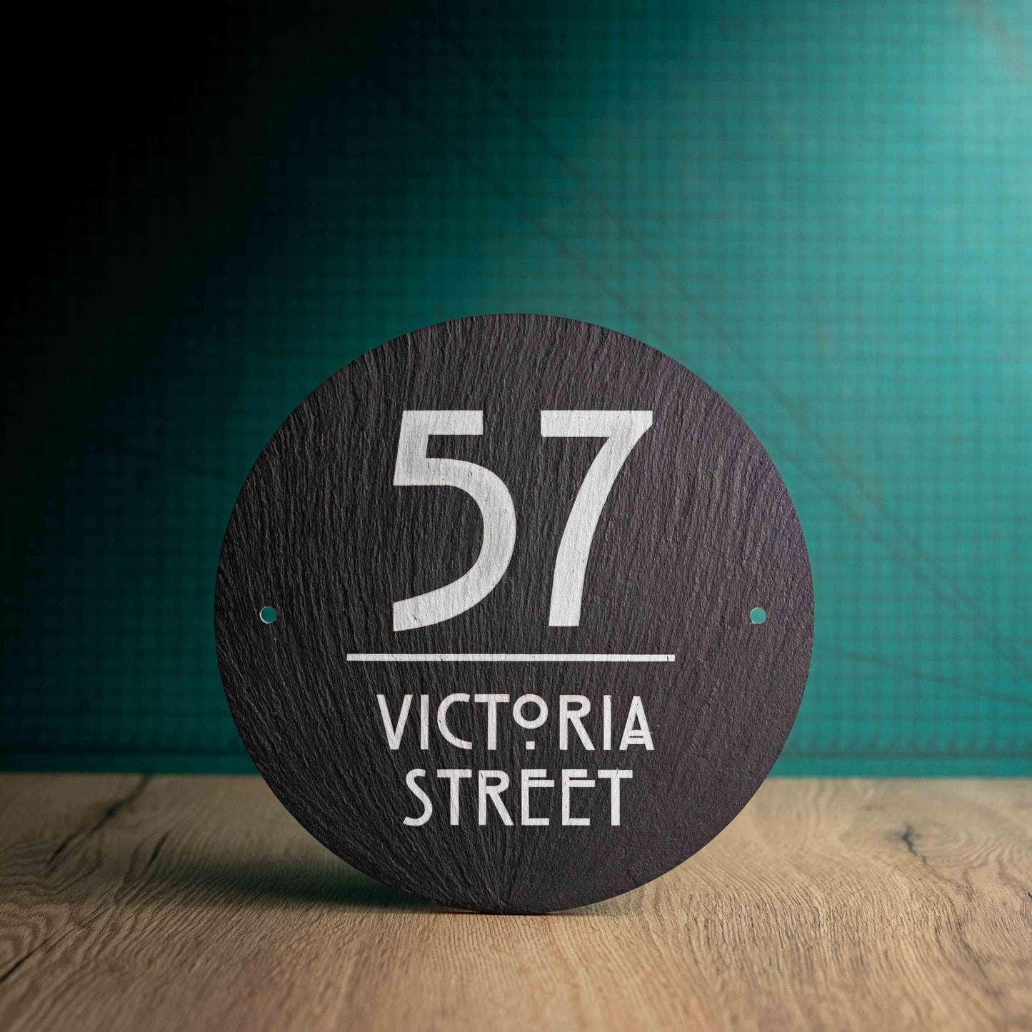A laser-engraved round slate house sign on a wooden surface and teal background. Circle 9"