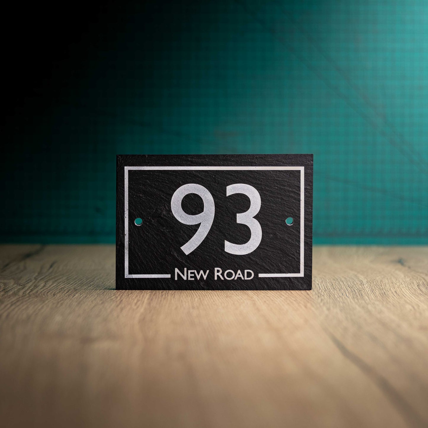 A laser-engraved rectangular slate house sign on a wooden surface and teal background. Rectangle 6" x 4"