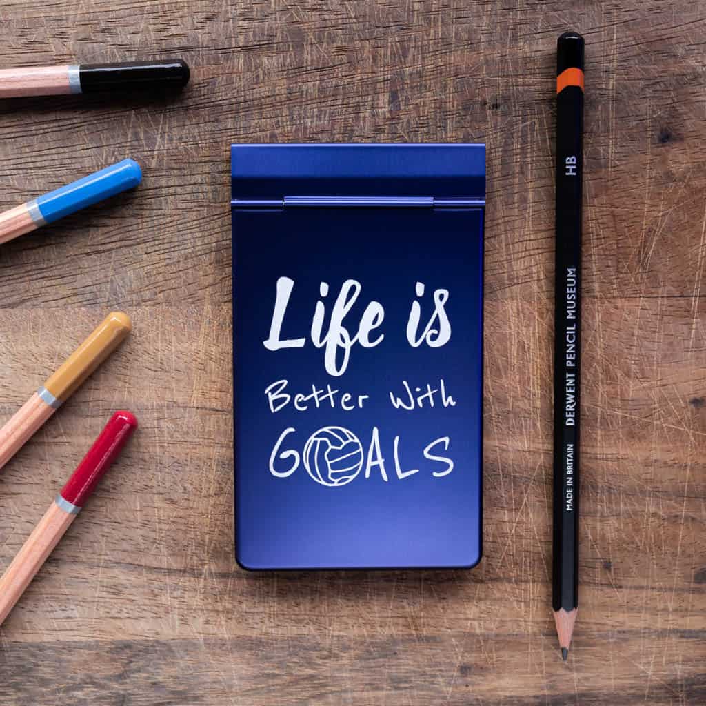 A notepad with a blue anodized metal cover with the phrase "Life is better with goals" laser marked onto the cover