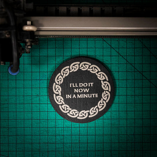 A laser engraved Welsh slate coaster with the words I'll do it now in a minute inside a celtic knot boarder