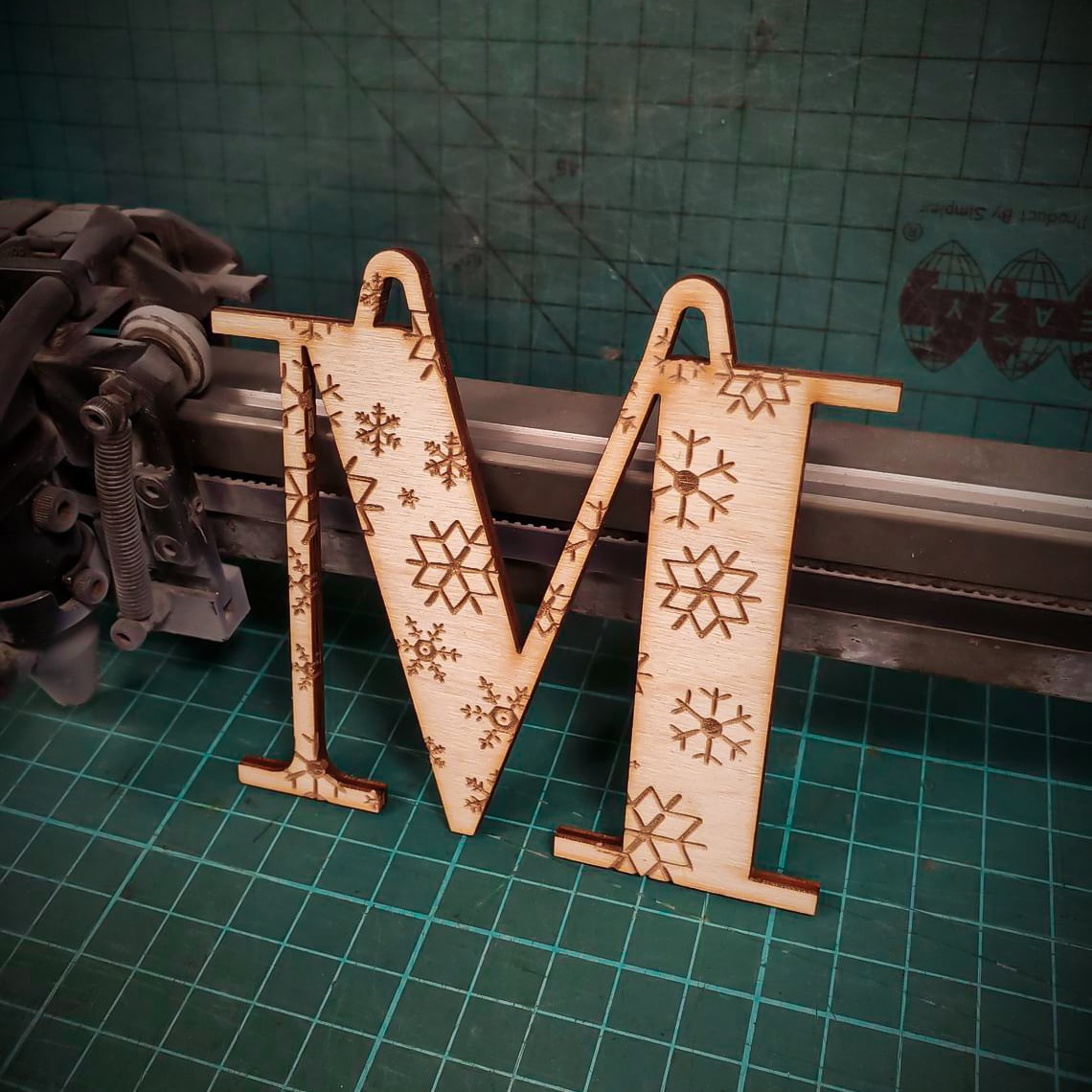 A wooden Christmas tree decoration laser cut into the shape of the letter "M" and engraved with snowflakes