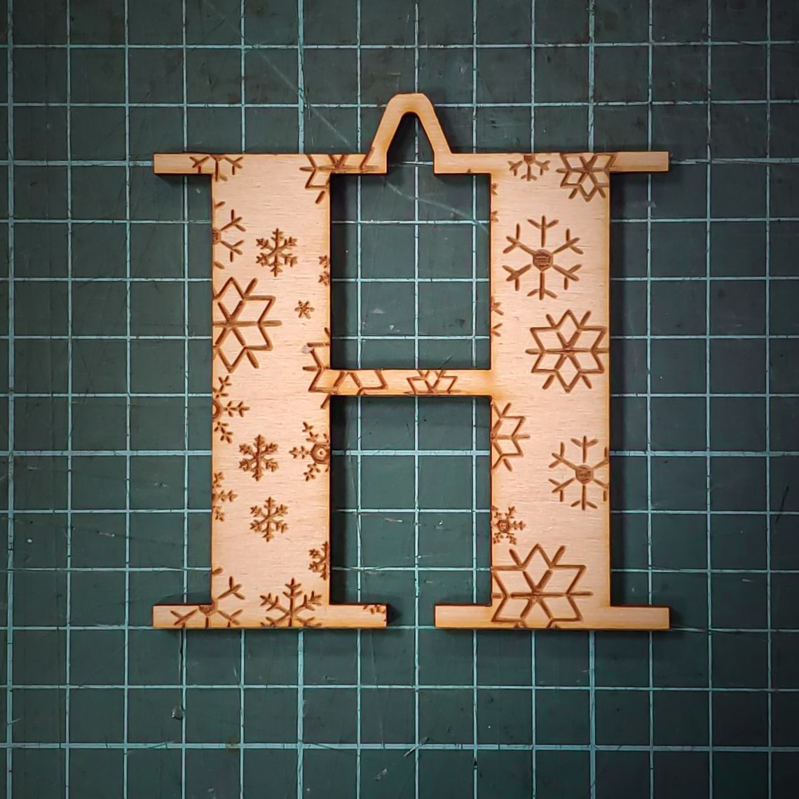 A wooden Christmas tree decoration laser cut into the shape of the letter "H" and engraved with snowflakes