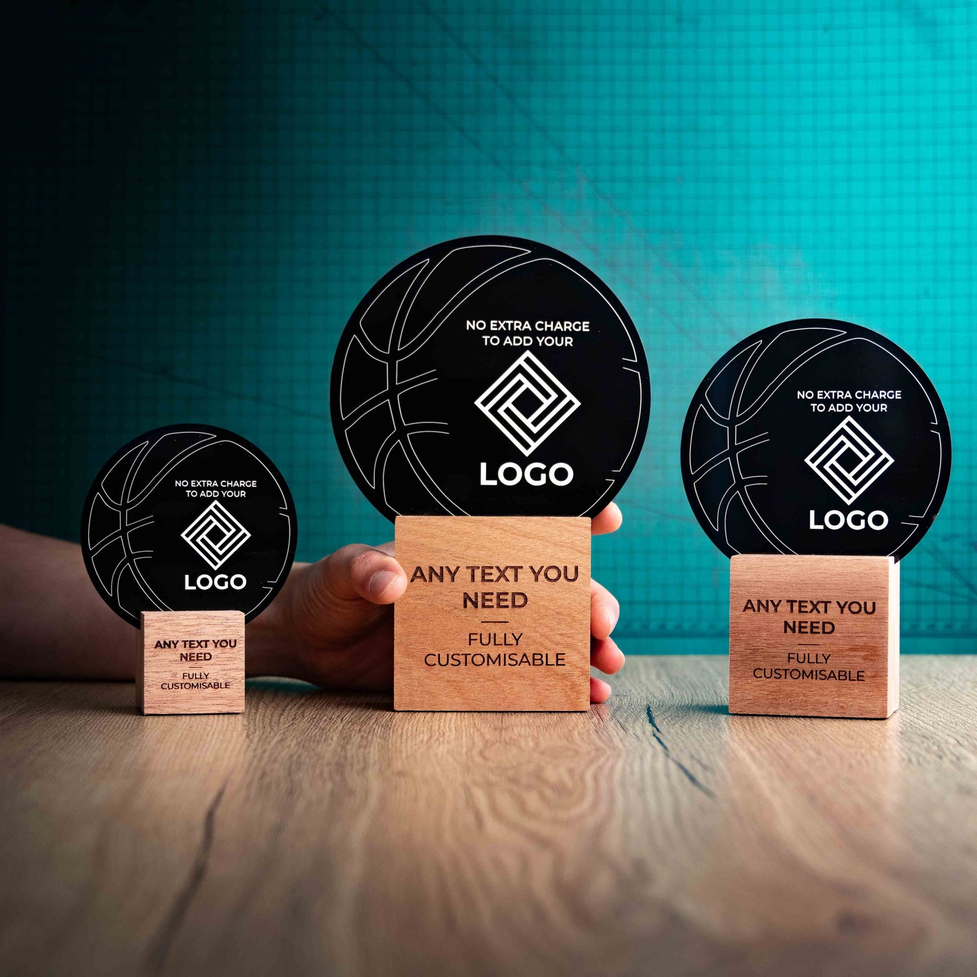 Three Basketball trophies with custom text and logos, on eco-friendly wooden bases, with a black Perspex Basketball mounted on top, featuring fully customisable areas for text and logos with a hand holding the large trophy to give a perspective of the size.