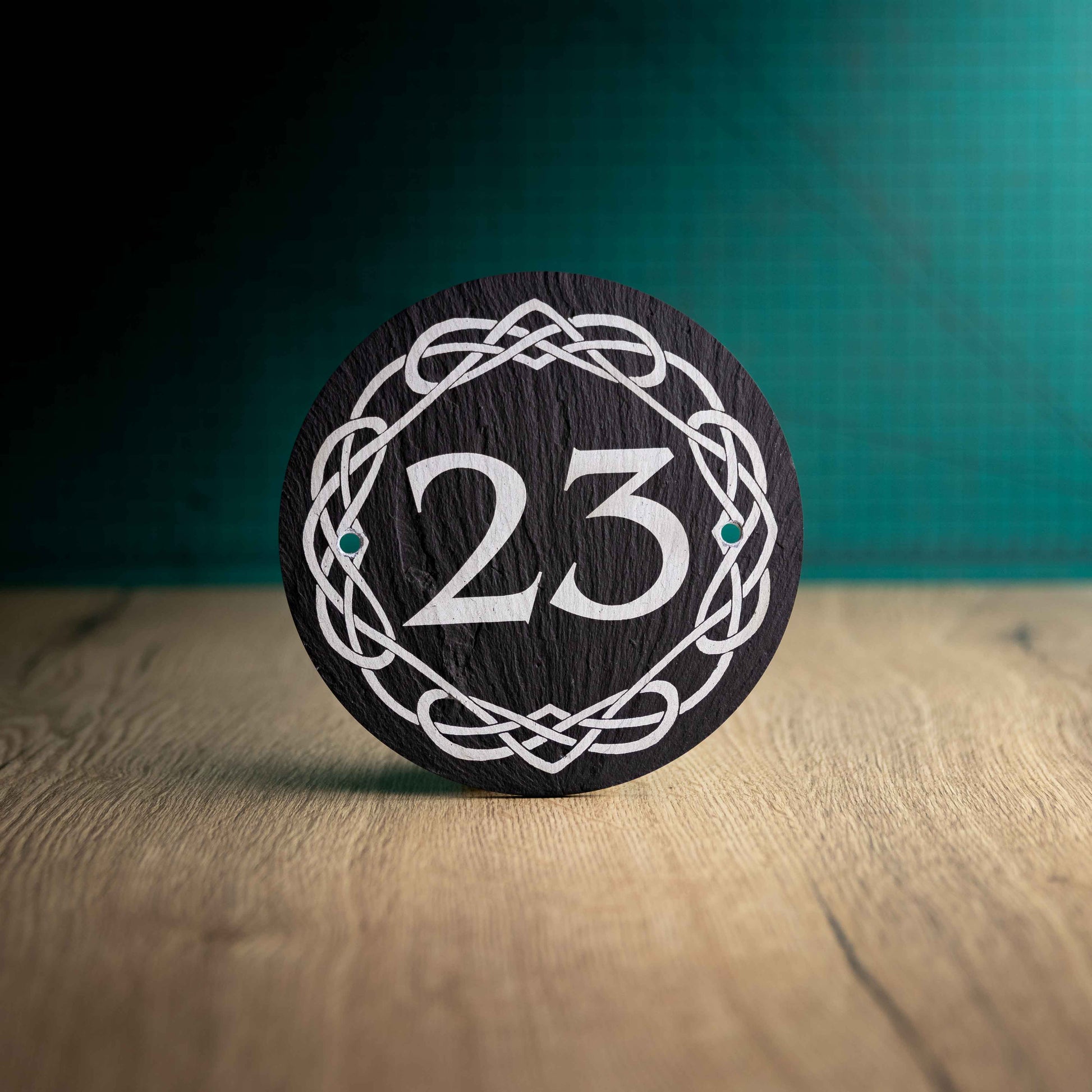 A laser-engraved round slate house sign with celtic border on a wooden surface and teal background. Circle 3.75", 6" or 9"