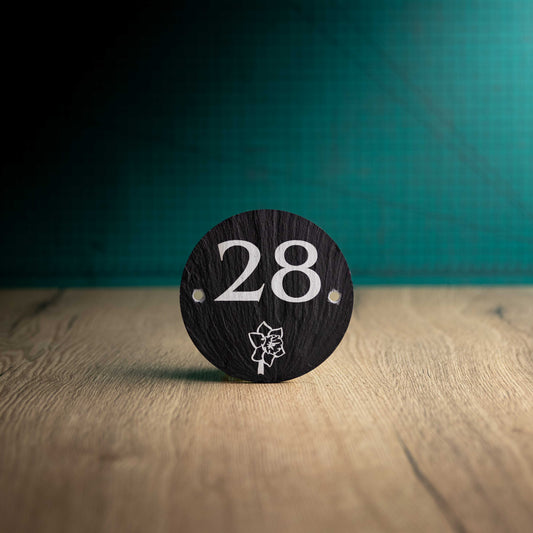 A laser-engraved round slate house sign on a wooden surface and teal background. Circle 3.75"