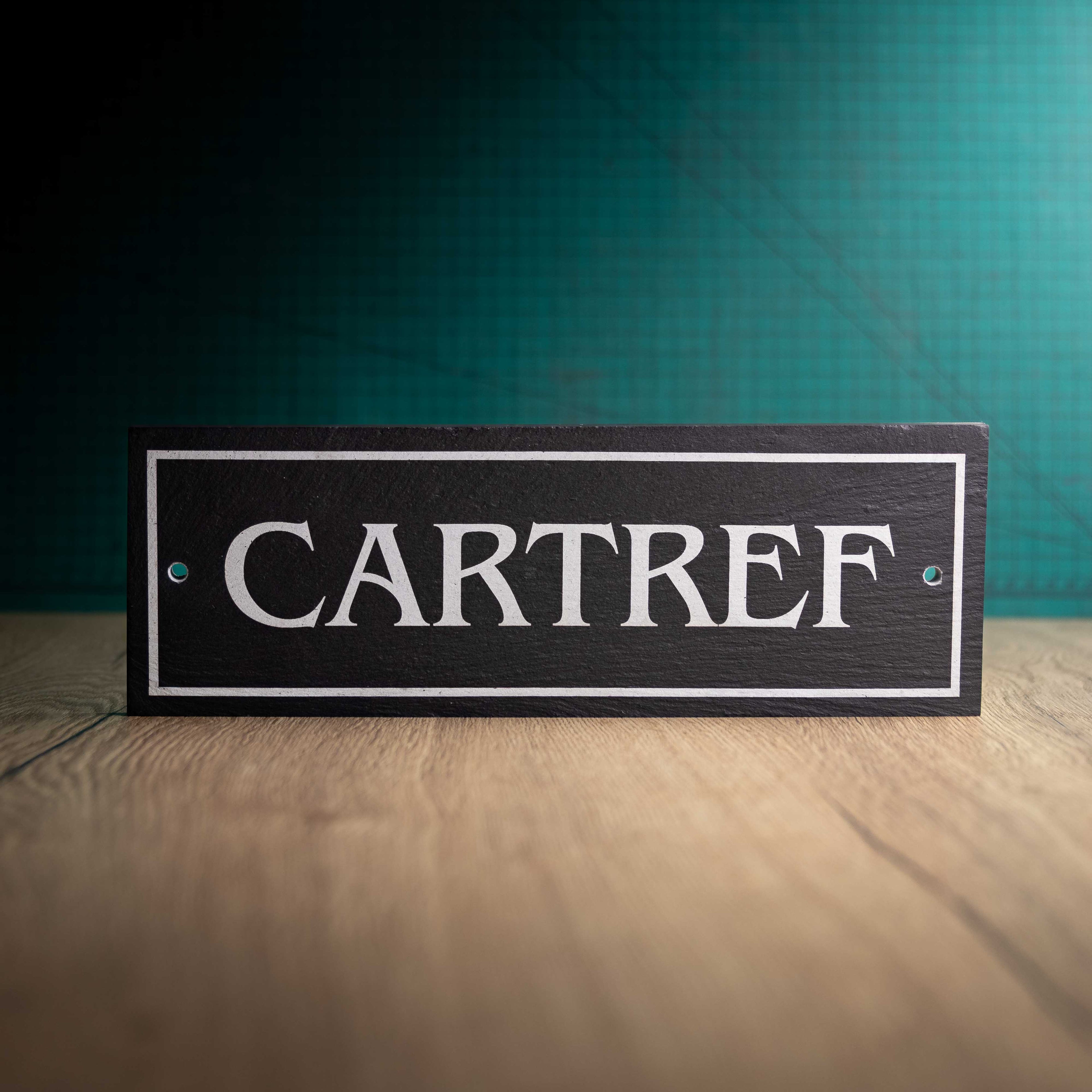 A laser-engraved slate house sign with 'CARTREF' on a wooden surface and teal background. Long Rectangle 12" x 4" x 0.5"