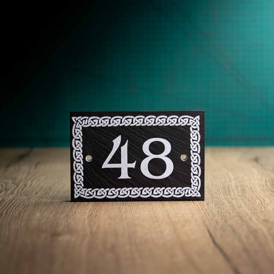 A laser-engraved slate house sign with a celtic border on a wooden surface and teal background. Rectangle 6" x 4"