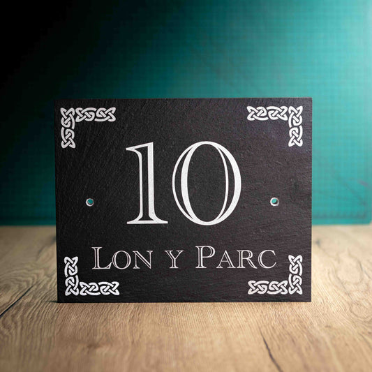 A laser-engraved slate house sign with a House Name, House Number and a Celtic Knot motif in each corner on a wooden surface and teal background. Large Rectangle 10" x 8" x 0.5"