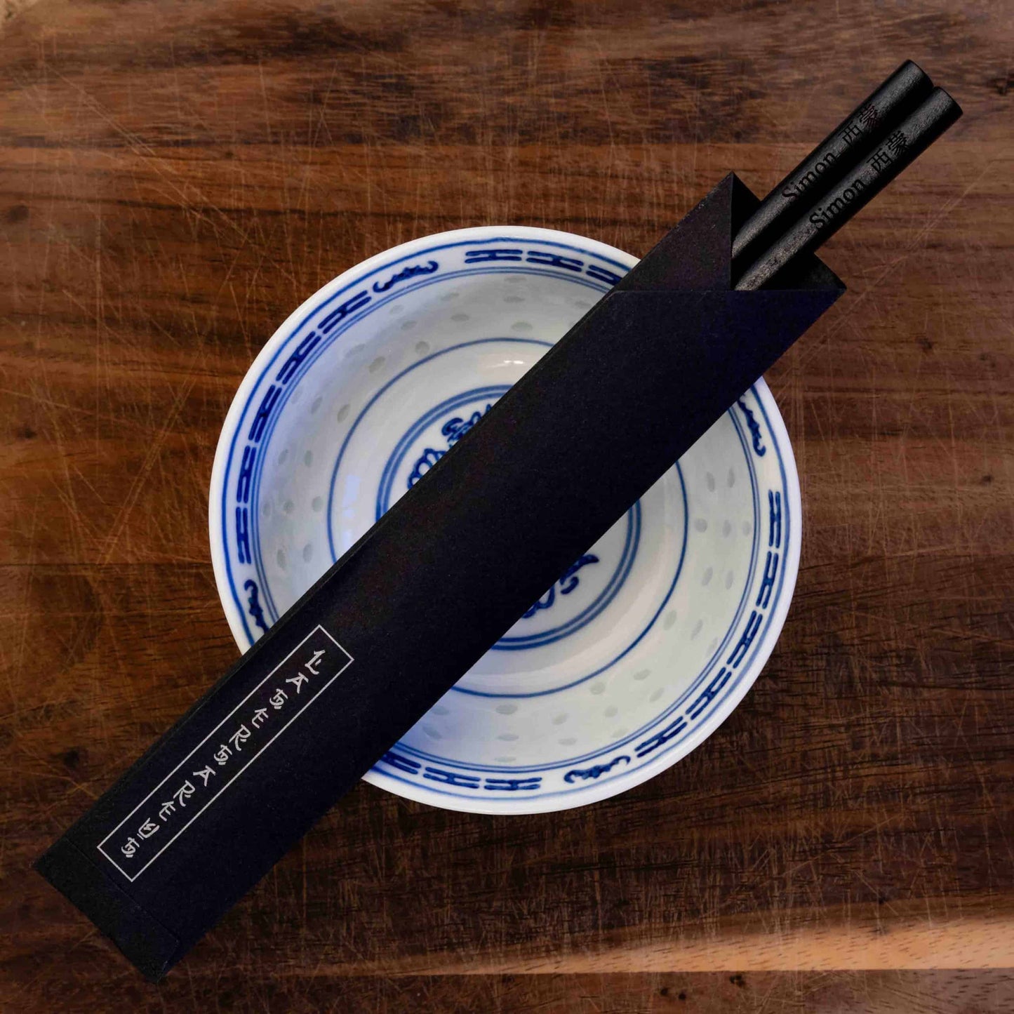 A pair of Japanese-style Chopsticks in a paper pouch, colour black wood. The chopsticks are laser engraved and personalised with either a name or a phrase in English and are translated into either Chinese or Japanese.
