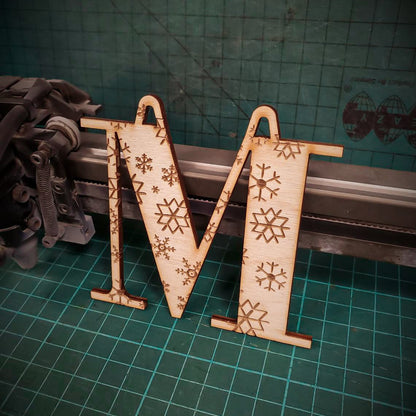 A wooden Christmas tree decoration laser cut into the shape of the letter "M" and engraved with snowflakes