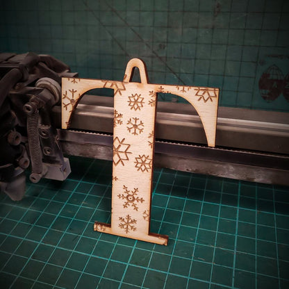 A wooden Christmas tree decoration laser cut into the shape of the letter "T" and engraved with snowflakes