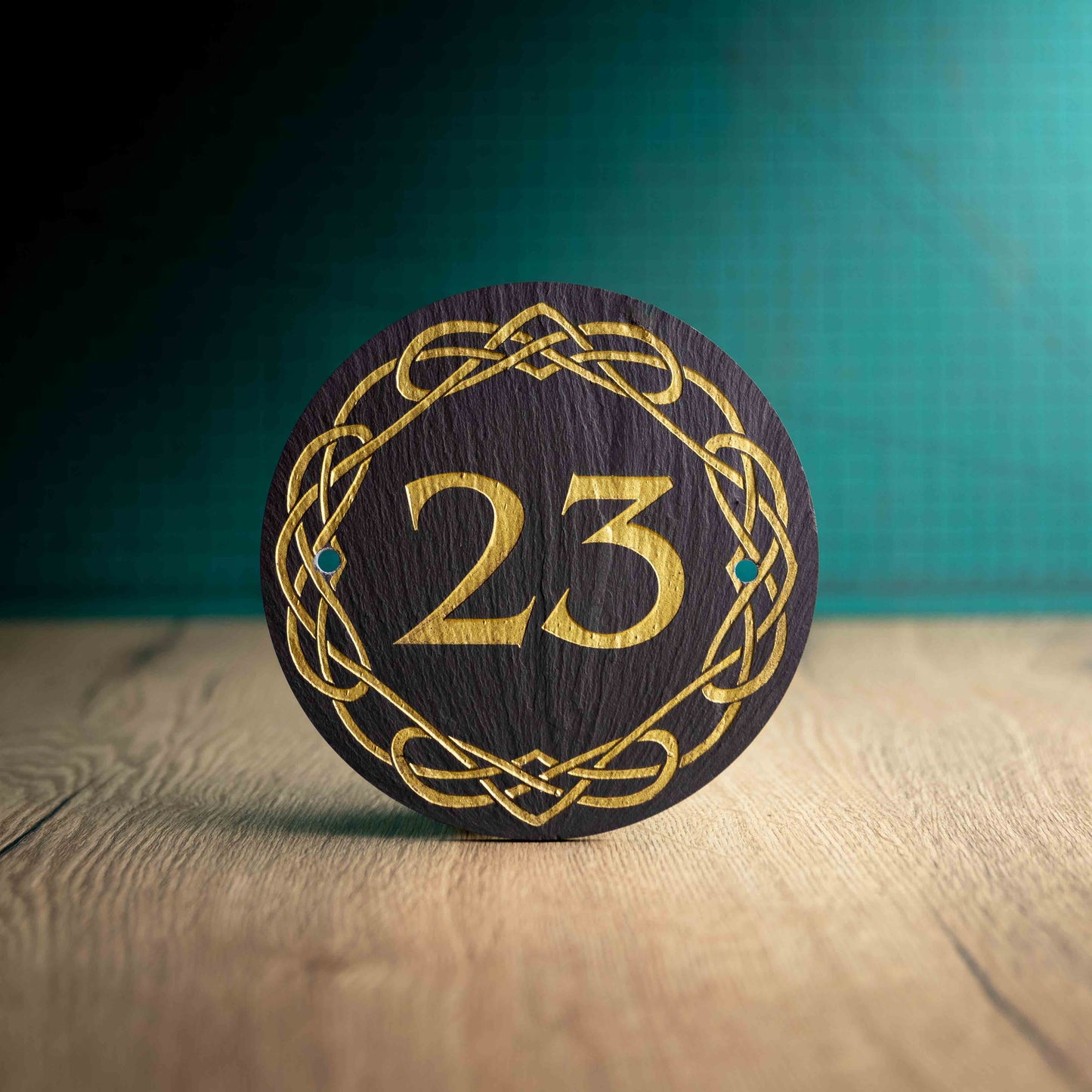 A laser-engraved, gold colour-filled, round slate house sign with a Celtic border on a wooden surface and teal background. Circle 3.75", 6" or 9"