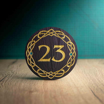 A laser-engraved, gold colour-filled, round slate house sign with a Celtic border on a wooden surface and teal background. Circle 3.75", 6" or 9"