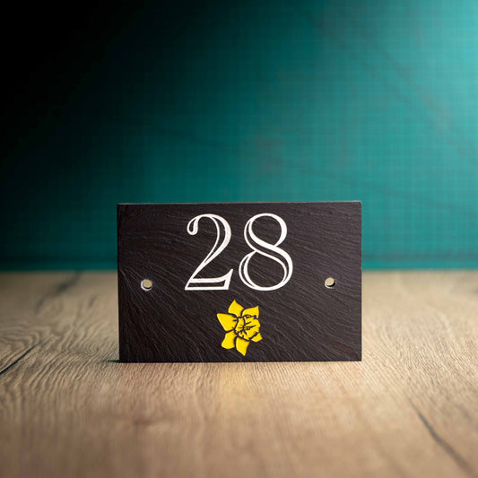 A laser-engraved colour-filled slate house sign with a Welsh Emblem on a wooden surface and teal background. Rectangle 6" x 4"