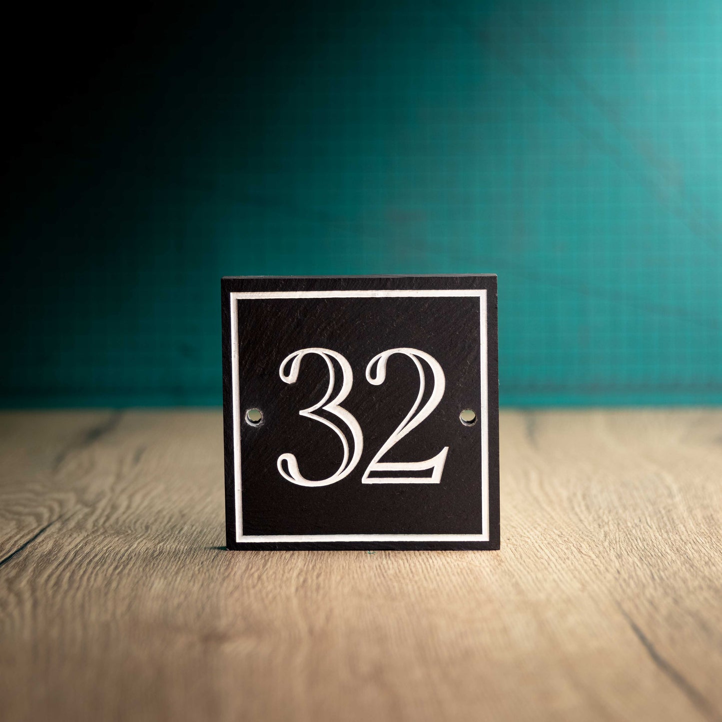 A laser-engraved square slate house sign on a wooden surface and teal background. Square 4.5" x 4.5"