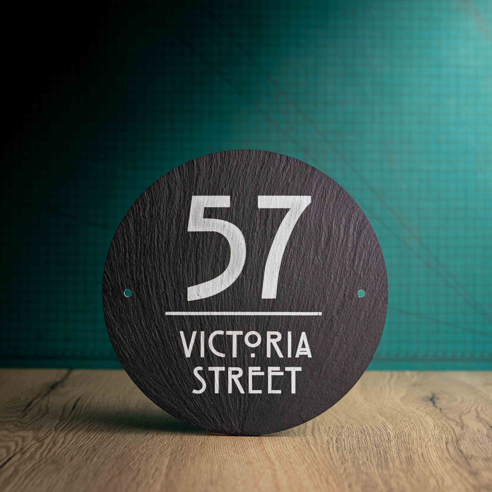 A laser-engraved round slate house sign on a wooden surface and teal background. Circle 9"