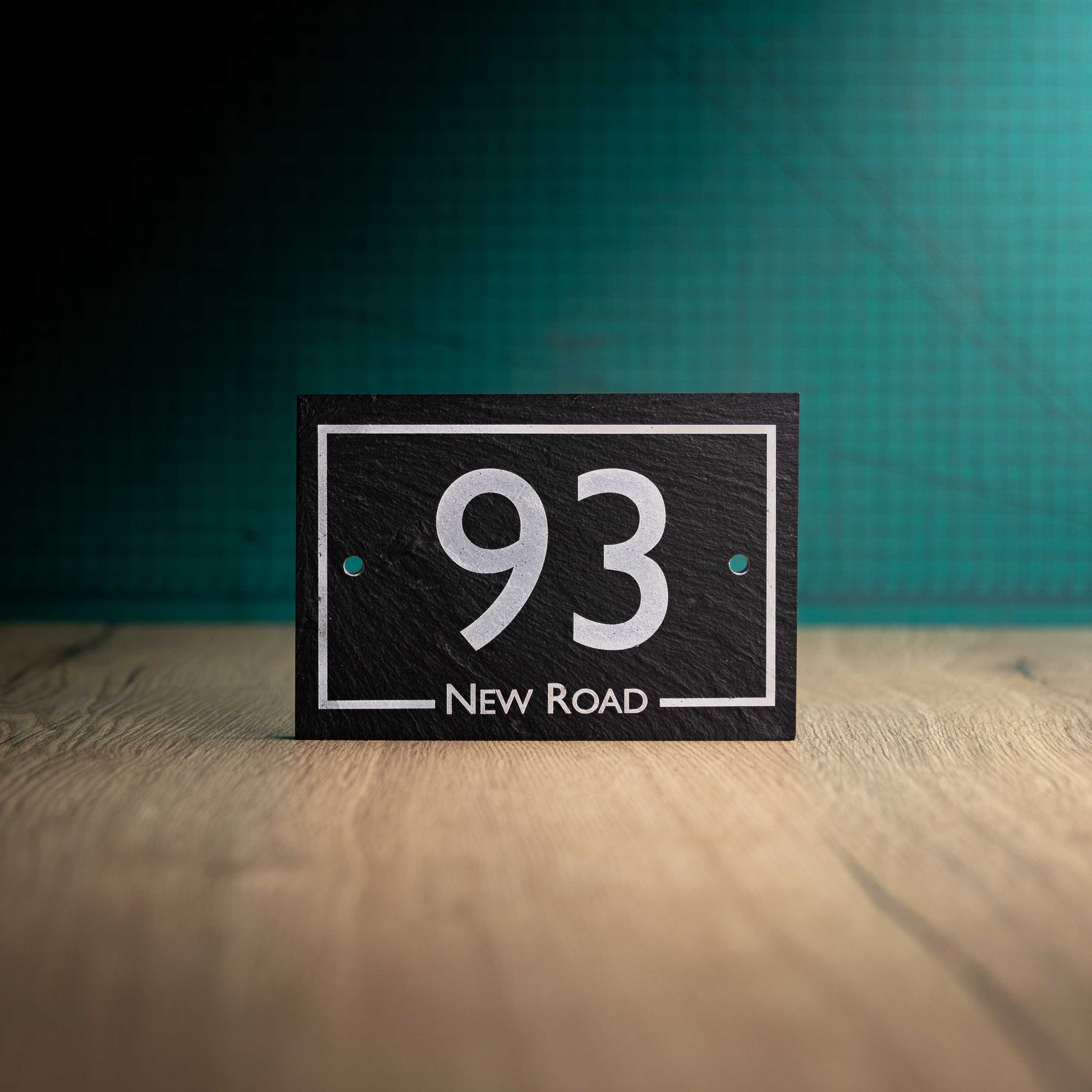 A laser-engraved rectangular slate house sign on a wooden surface and teal background. Rectangle 6" x 4"