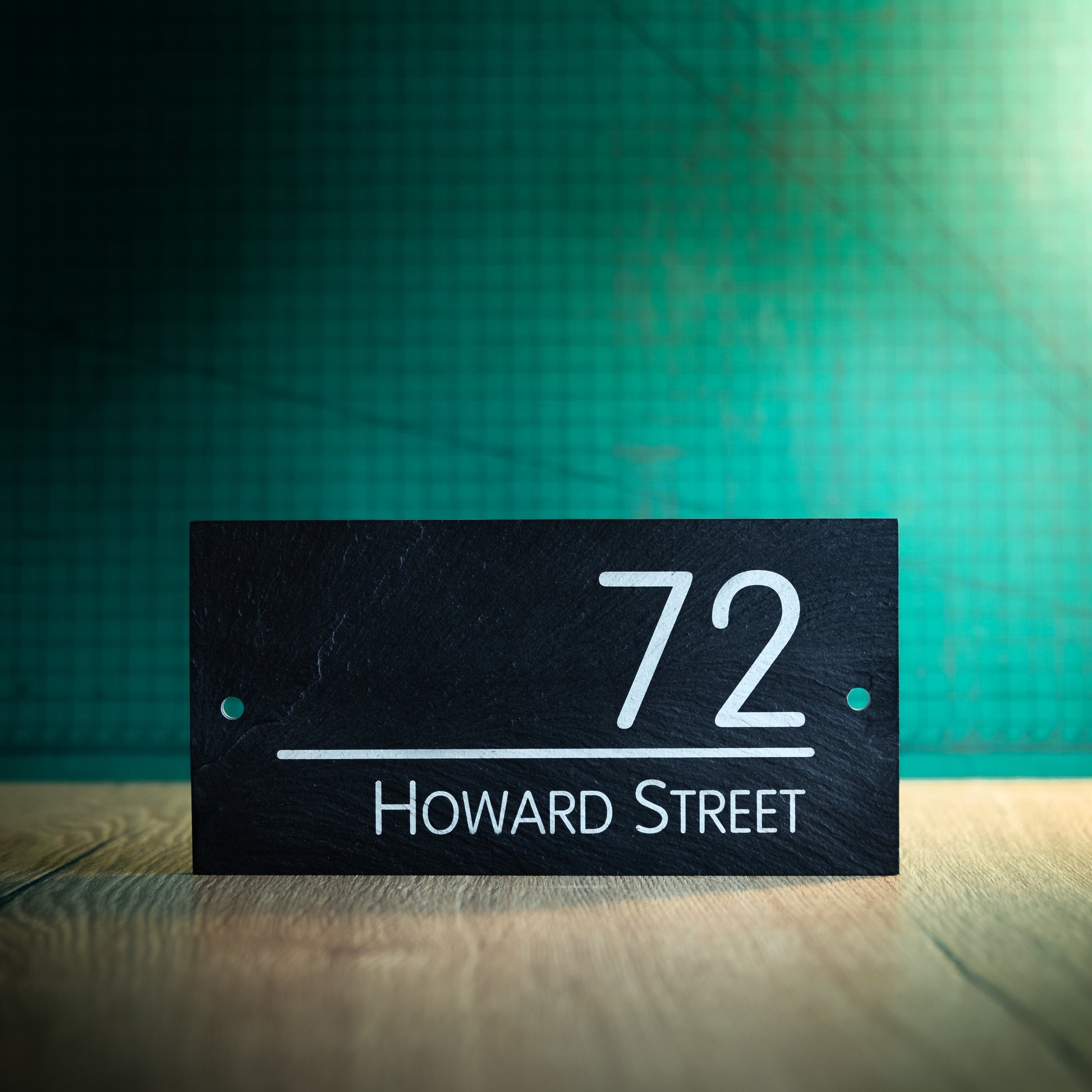 A laser-engraved rectangular slate house sign on a wooden surface and teal background. Rectangle 8" x 4"