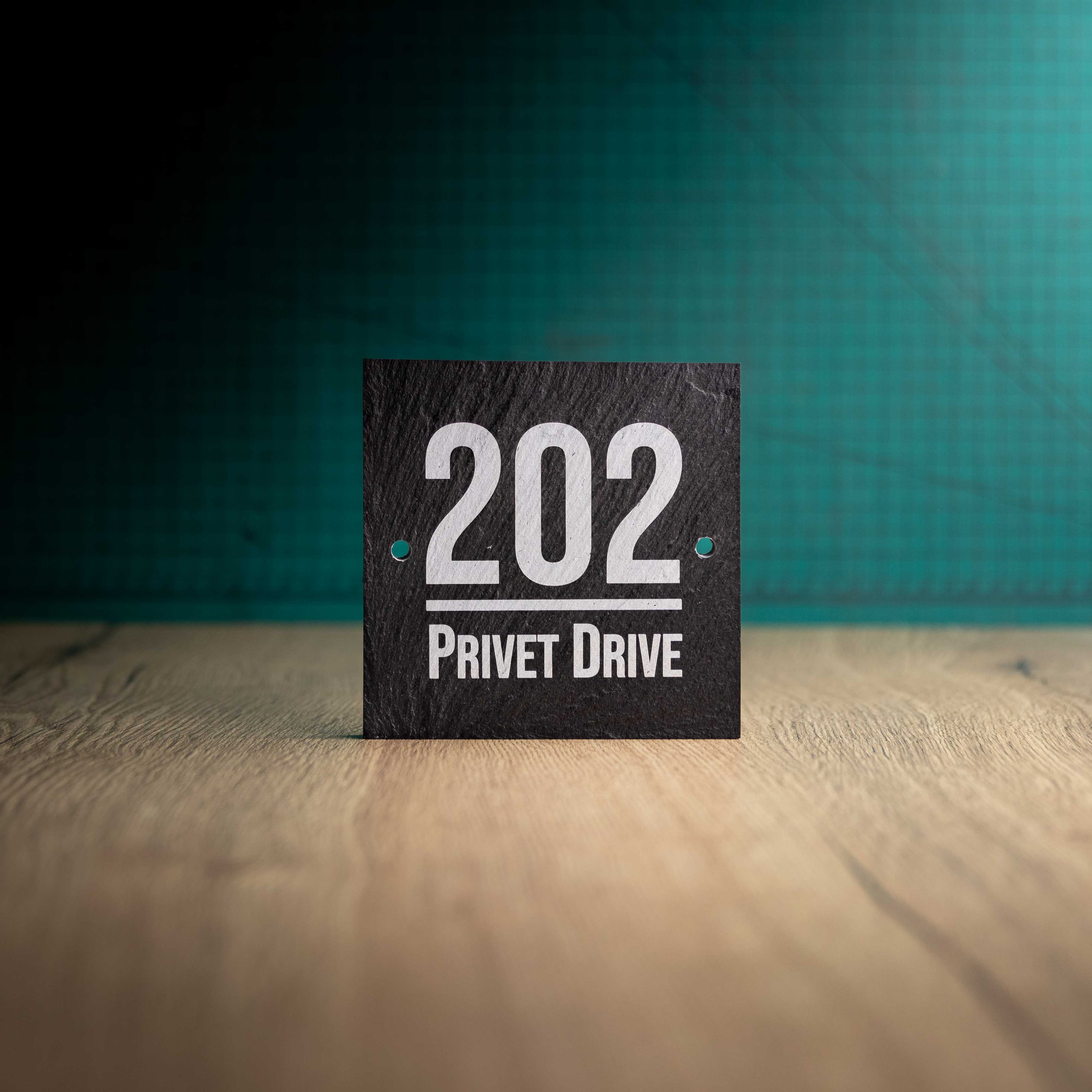 A laser-engraved square slate house sign on a wooden surface and teal background. Square 4.5" x 4.5"