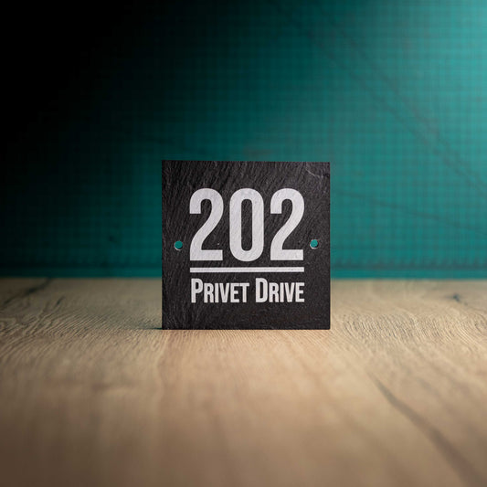 A laser-engraved square slate house sign on a wooden surface and teal background. Square 4.5" x 4.5"