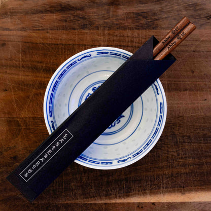 A pair of Japanese-style Chopsticks in a paper pouch, colour dark wood. The chopsticks are laser engraved and personalised with either a name or a phrase in English and are translated into either Chinese or Japanese.
