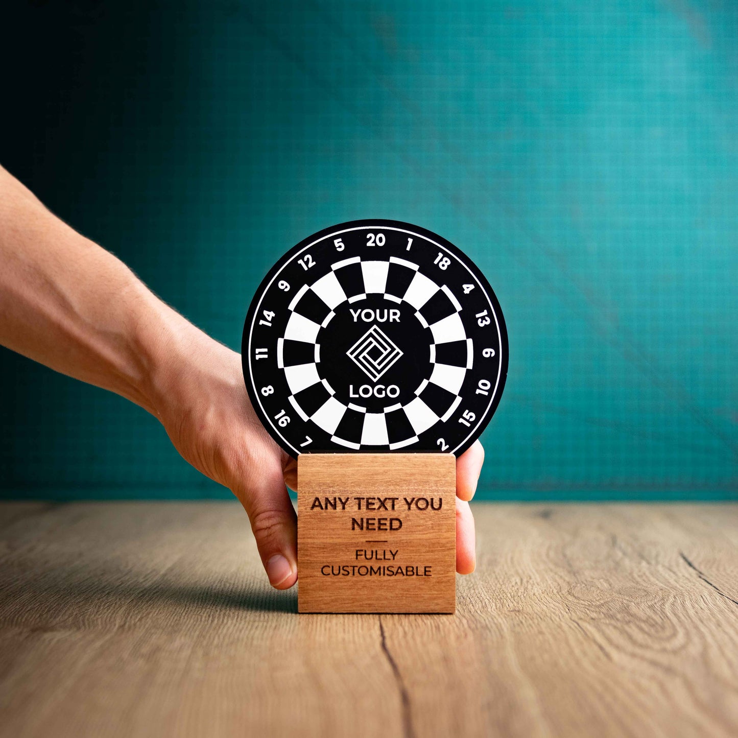 A hand holding a medium darts trophy with custom text and logos, on an eco-friendly wooden base, with a black Perspex dart board mounted on top, featuring fully customisable areas for text and logos.