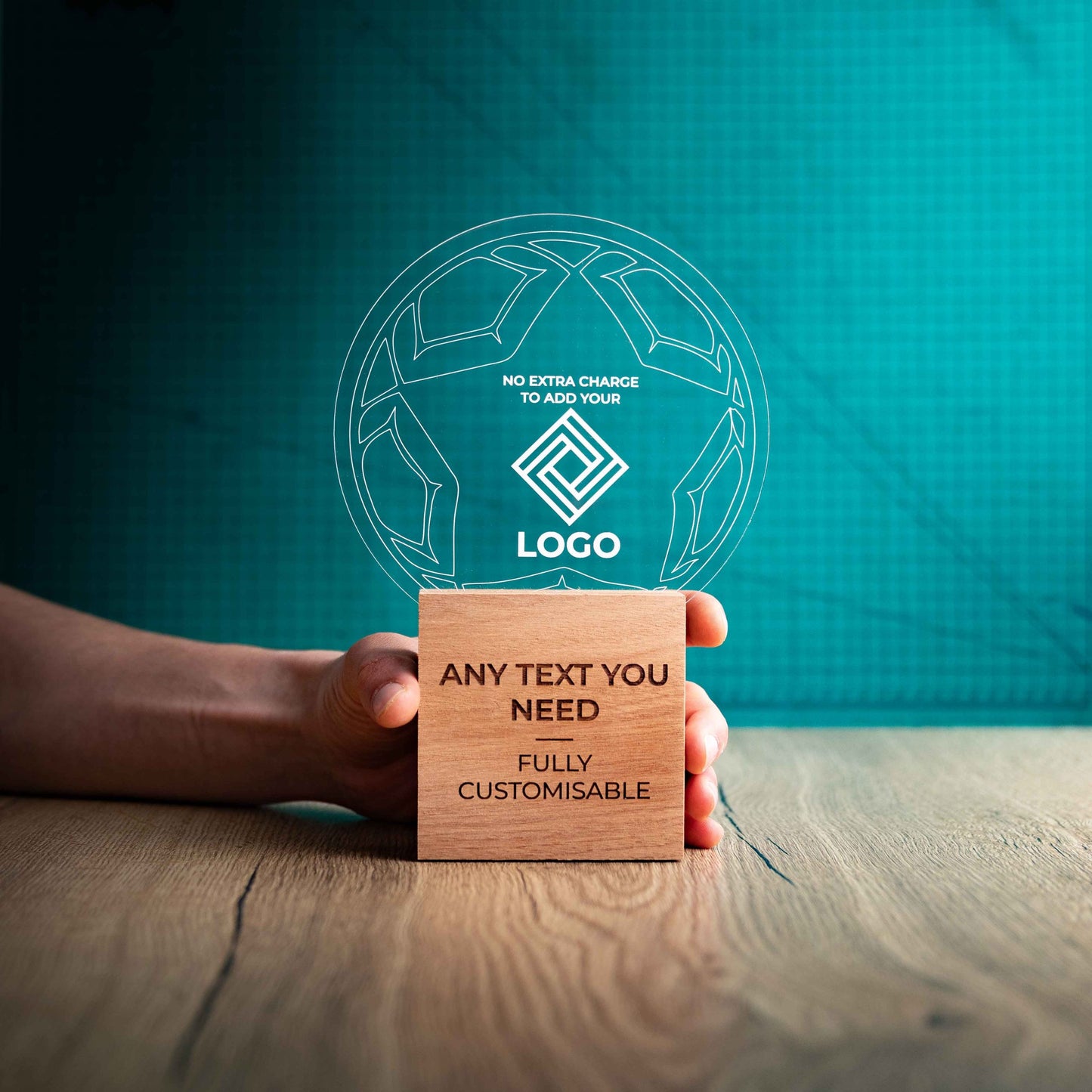 A hand holding a large football trophy with custom text and logos, on an eco-friendly wooden base, with a clear Perspex football mounted on top, featuring fully customisable areas for text and logos.