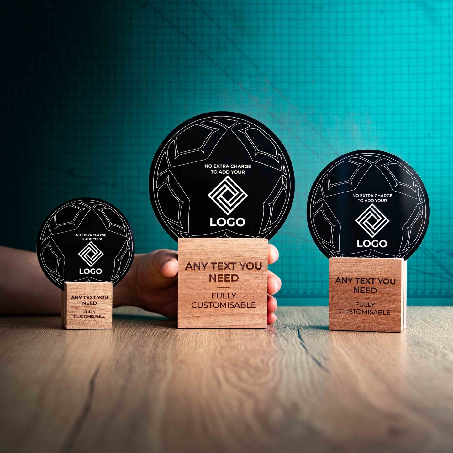 A hand holding a large football trophy, with small and medium versions on either side, custom text and logos, eco-friendly wooden bases, and a clear Perspex football mounted on top, with fully customisable areas featuring text and logos.