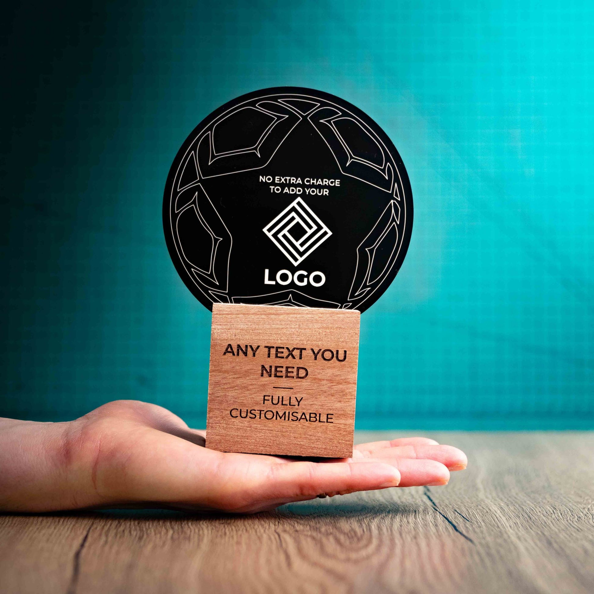 A hand holding a medium football trophy with custom text and logos, on an eco-friendly wooden base, with a black Perspex football mounted on top, featuring fully customisable areas for text and logos.