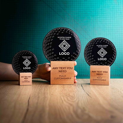Three golf award trophies with custom text and logos, on eco-friendly wooden bases, with a black Perspex golf ball mounted on top, with fully customisable areas featuring fully customisable areas for text and logos. A hand is present to show the size of the awards. 