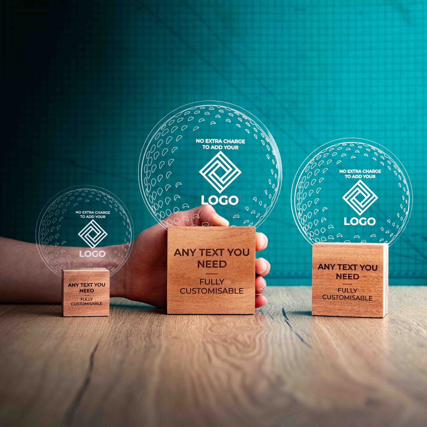 Three golf award trophies with custom text and logos, on eco-friendly wooden bases, with a clear Perspex golf ball mounted on top, with fully customisable areas featuring fully customisable areas for text and logos. A hand is present to show the size of the awards. 