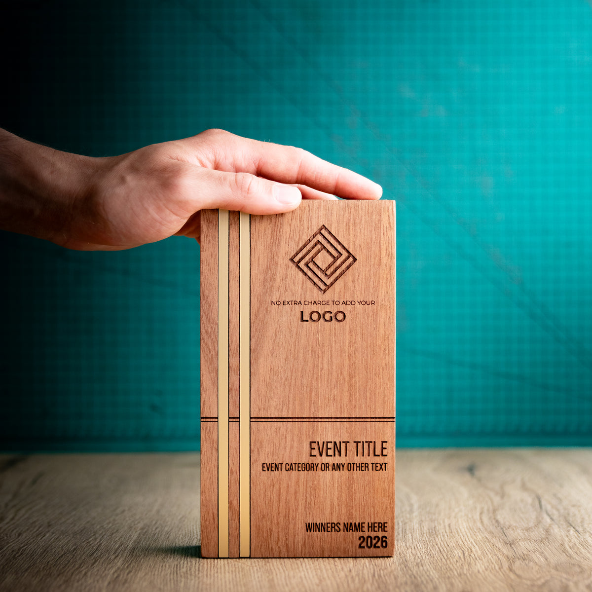 Large rectangular Kingdom wooden trophy standing upright with a hand resting across the top for scale. Two gold metallic inlays run the full height of the left side, with engraved logo, event and winner details on the front.