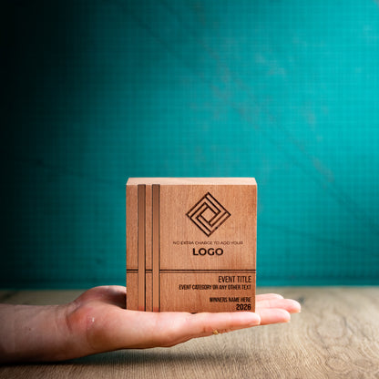 Small square Kingdom wooden trophy resting on an open hand for scale, with two bronze-toned metallic inlays running vertically along the left side and engraved logo, event and winner details on the front.