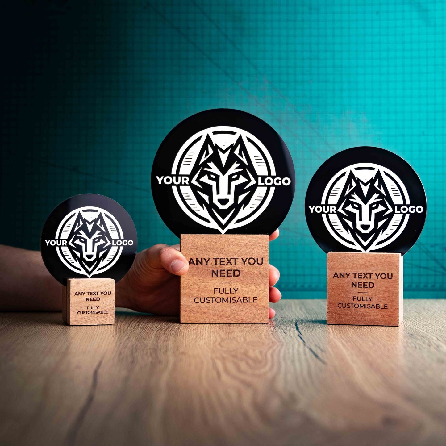 Three Legacy trophies with custom text and logos, on eco-friendly wooden bases, with a black Perspex circle mounted on top, featuring fully customisable areas for text and logos with a hand holding the large trophy to give a perspective of the size.