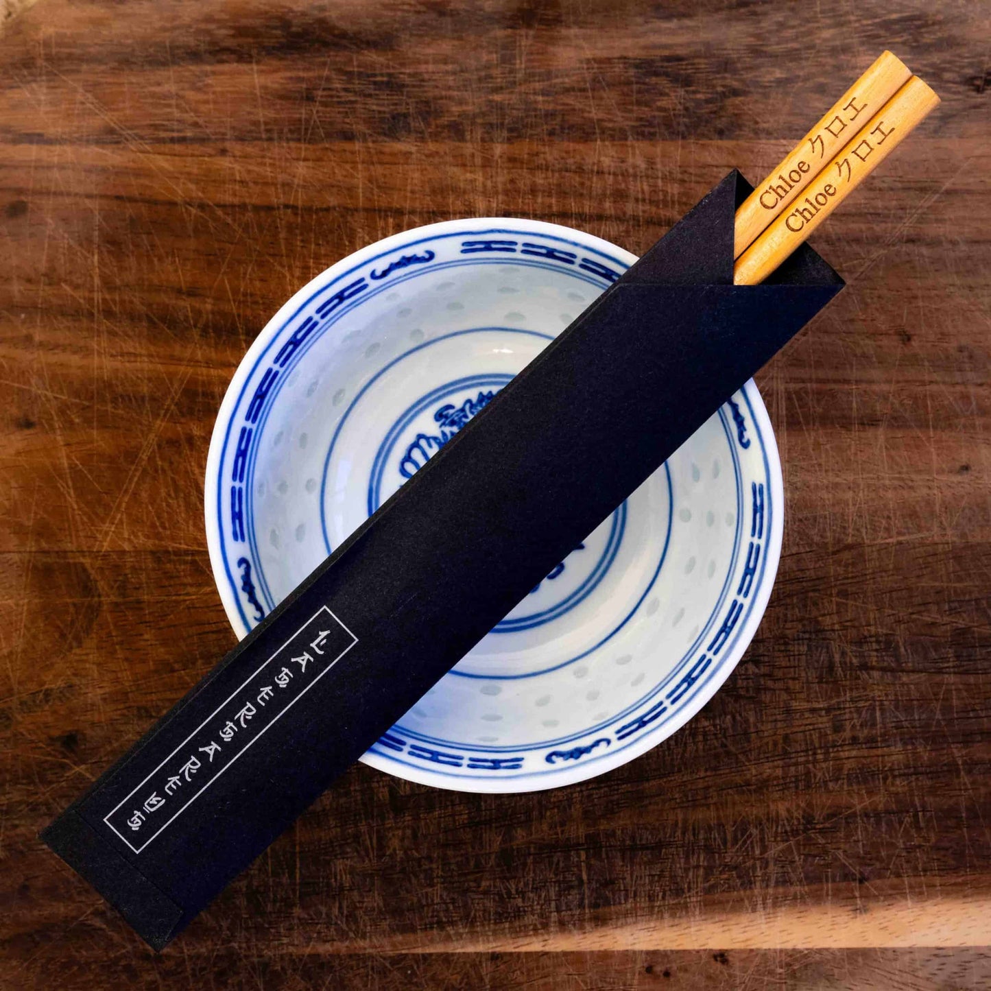 A pair of Japanese-style Chopsticks in a paper pouch, colour light wood. The chopsticks are laser engraved and personalised with either a name or a phrase in English and are translated into either Chinese or Japanese.