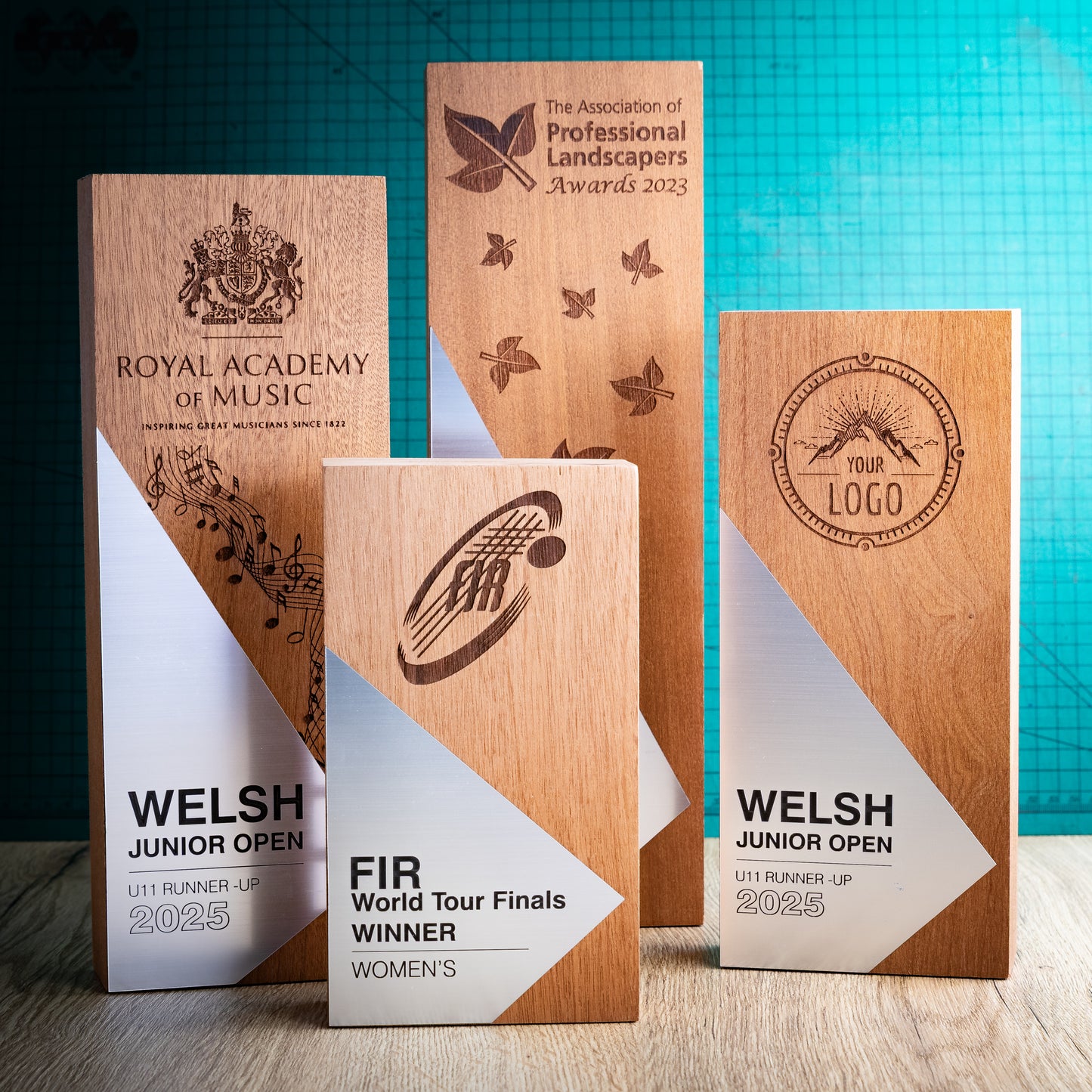 Four engraved wooden perpetual awards are displayed on a tabletop against a teal grid backdrop. The trophies feature natural wood grain, white and silver geometric panels, and dark brown laser engravings for various sports and organisation names, including the Royal Academy of Music, Welsh Junior Open, and FIR World Tour Finals.