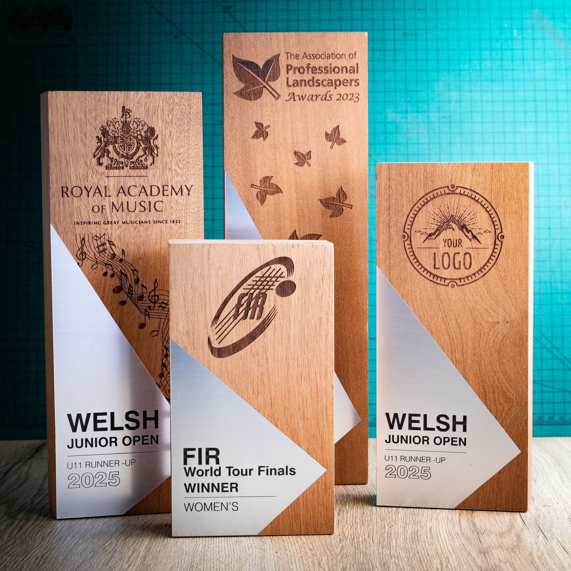 Four engraved wooden perpetual awards are displayed on a tabletop against a teal grid backdrop. The trophies feature natural wood grain, white and silver geometric panels, and dark brown laser engravings for various sports and organisation names, including the Royal Academy of Music, Welsh Junior Open, and FIR World Tour Finals.