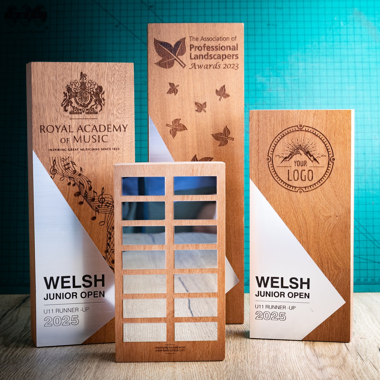 Four engraved wooden perpetual awards are displayed on a tabletop against a teal grid backdrop. The trophies feature natural wood grain, white and silver geometric panels, and dark brown laser engravings for various sports and organisation names, including the Royal Academy of Music, Welsh Junior Open, and Professional Landscapers Awards.