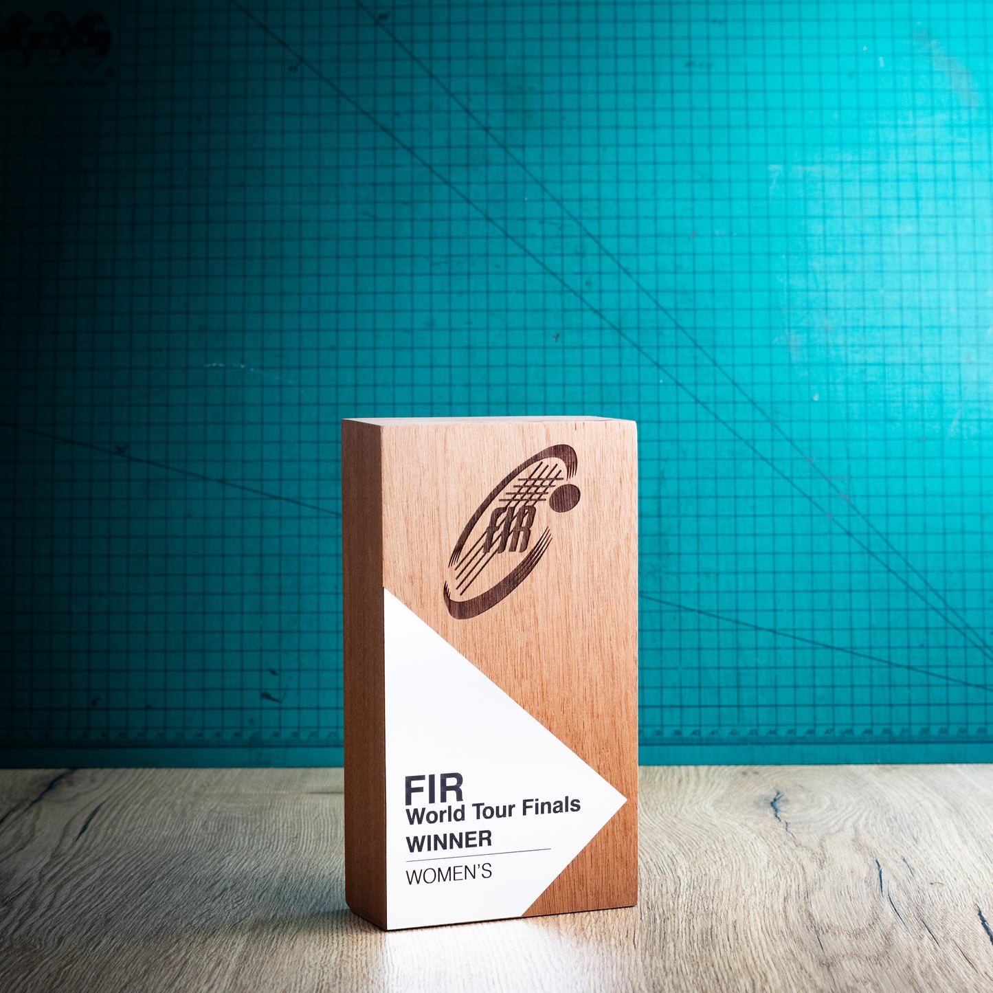 A wooden perpetual award trophy standing upright on a light wood surface, with a teal grid backdrop behind it. The front of the plaque is white and printed with the text 'FIR World Tour Finals Winner Women' and a dark engraved logo.