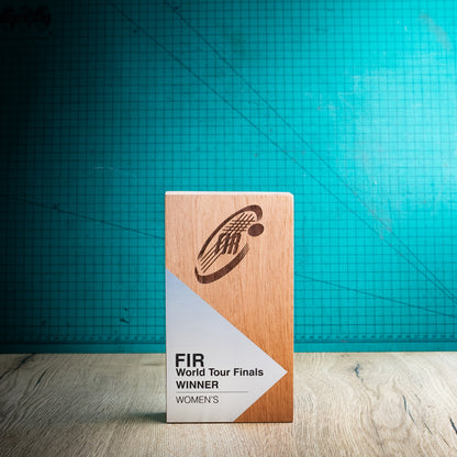 A wooden perpetual award trophy standing upright on a light wood surface, with a teal grid backdrop behind it. The front of the plaque is white and printed with the text 'FIR World Tour Finals Winner Women' and a dark engraved logo.