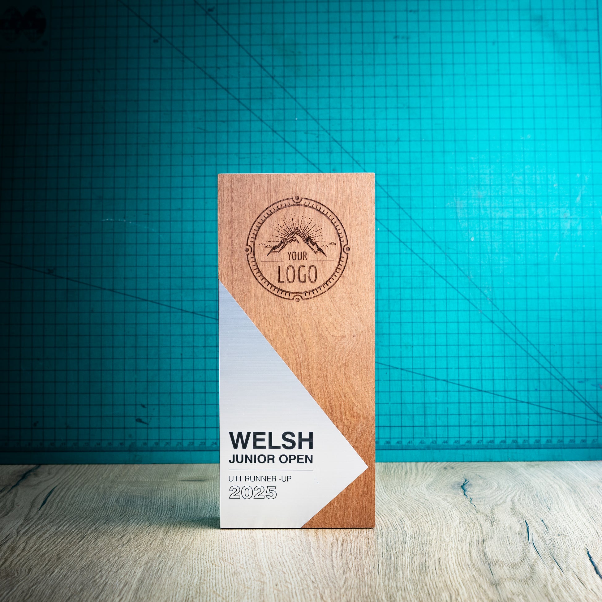 A rectangular wooden perpetual award trophy for the Welsh Junior Open 2025, standing upright on a light wood surface against a teal grid background.