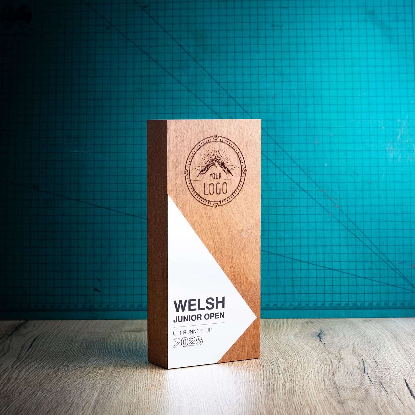 A rectangular wooden perpetual award trophy for the Welsh Junior Open 2025, standing upright on a light wood surface against a teal grid background.
