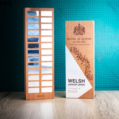 Two wooden perpetual award trophies, standing on a light wood floor against a teal grid background. One trophy features the perpetual award plaques with enough room for 26 years' worth of winners, and the other is engraved with the Royal Academy of Music crest and musical notation on a light wood grain surface.