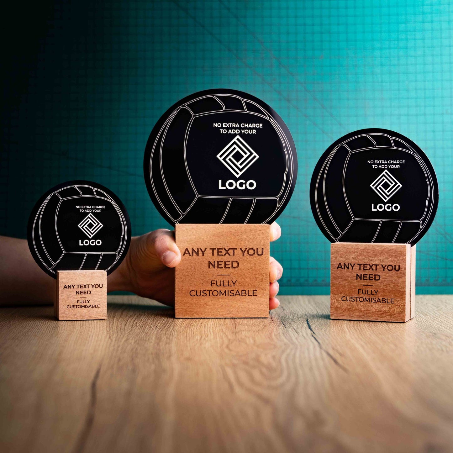 Three Netball trophies with custom text and logos, on eco-friendly wooden bases, with a black Perspex Netball mounted on top, featuring fully customisable areas for text and logos with a hand holding the large trophy to give a perspective of the size.