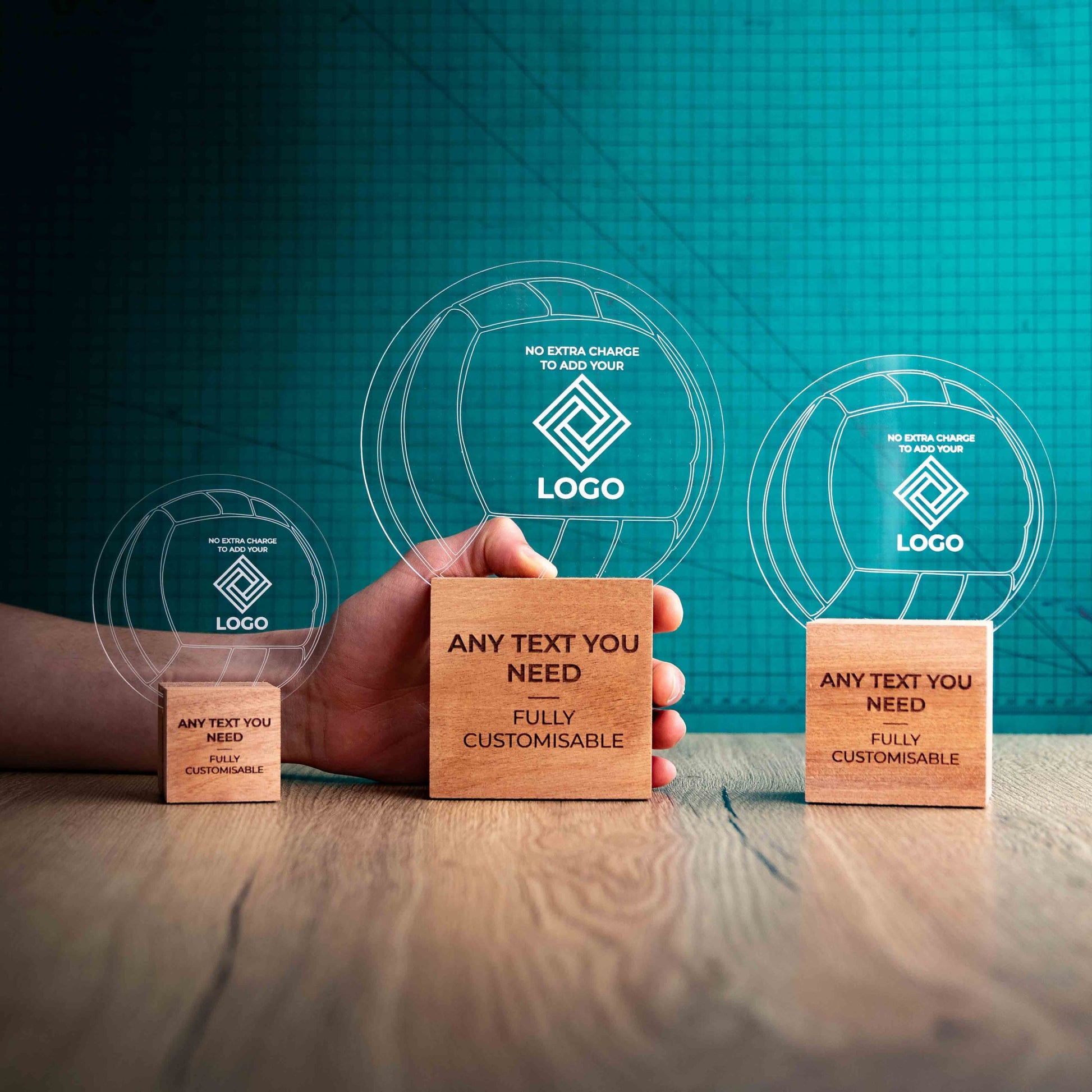 Three Netball trophies with custom text and logos, on eco-friendly wooden bases, with a clear Perspex Netball mounted on top, featuring fully customisable areas for text and logos with a hand holding the large trophy to give a perspective of the size.