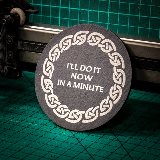 A laser engraved Welsh slate coaster with the words I'll do it now in a minute inside a celtic knot boarder