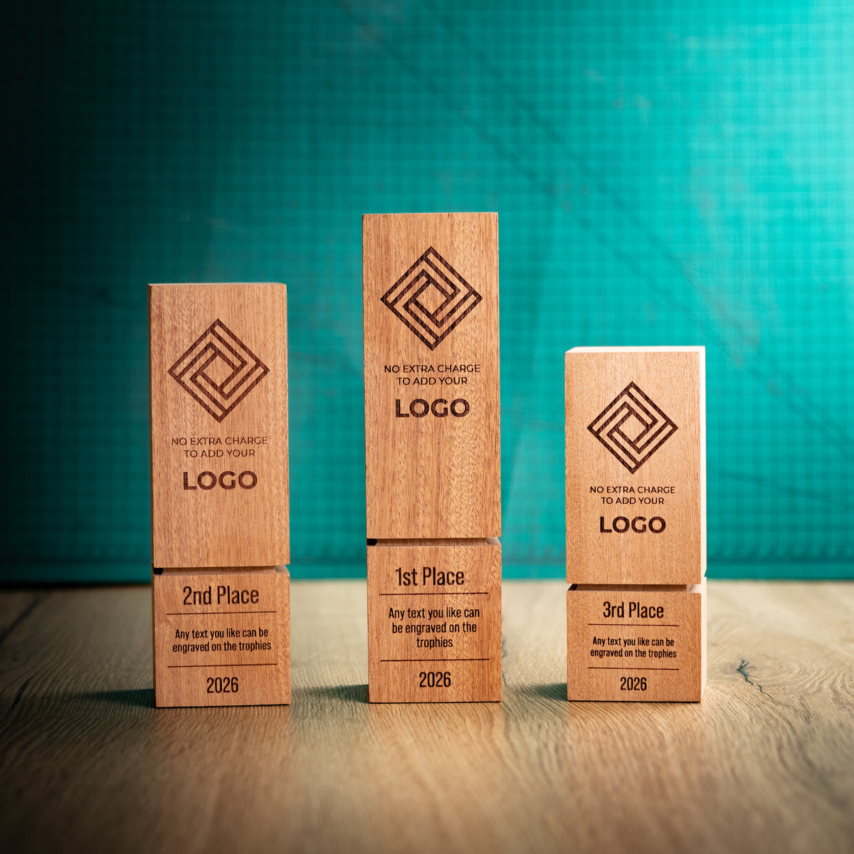 Three wooden block trophies of different heights with a distinctive recessed groove near their bases, standing on a wooden surface against a textured teal background. Each block is engraved with a geometric logo, placeholder text, and a place rank: 1st, 2nd, and 3rd.