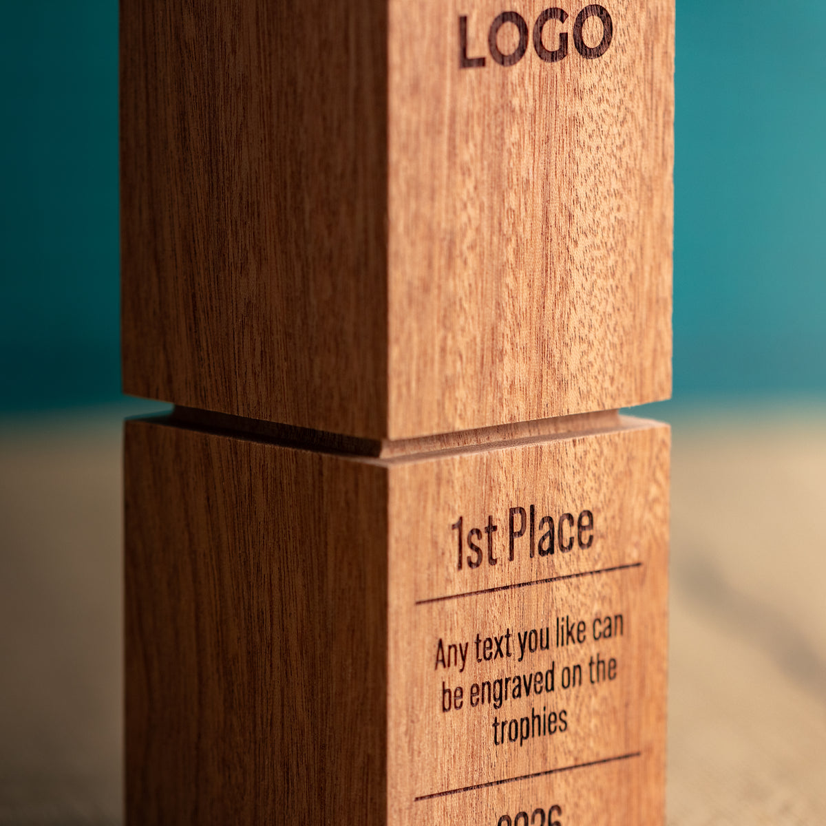 Close-up of a wooden trophy block with engraved text reading 'LOGO' and '1st Place' on a warm wood surface against a teal background.